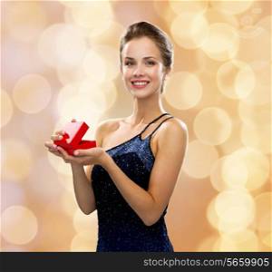 holidays, presents, luxury and happiness concept - smiling woman in dress holding red gift box over beige lights background