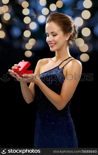 holidays, presents, luxury and happiness concept - smiling woman in dress holding red gift box night lights background