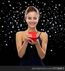 holidays, presents, luxury and happiness concept - smiling woman in dress holding red gift box over black snowy background