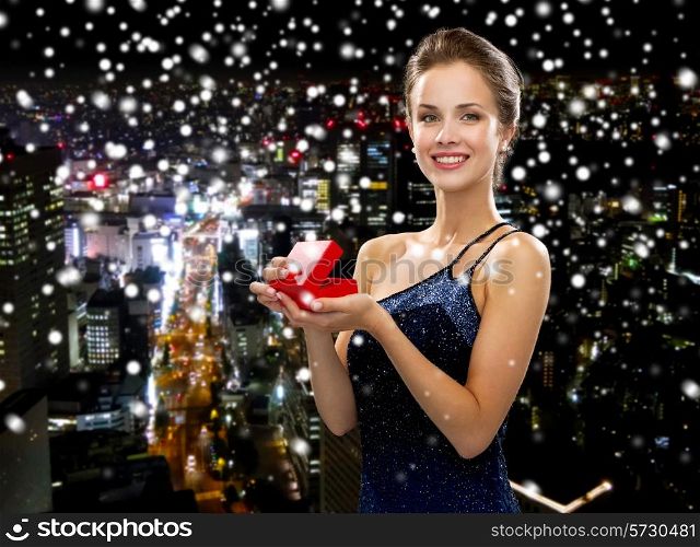 holidays, presents, luxury and happiness concept - smiling woman in dress holding red gift box over snowy night city background