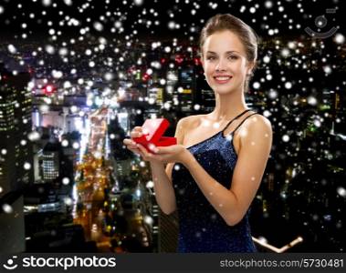 holidays, presents, luxury and happiness concept - smiling woman in dress holding red gift box over snowy night city background