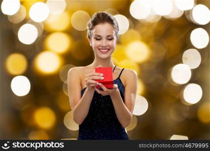 holidays, presents, luxury and people concept - smiling woman in dress holding red gift box over lights background