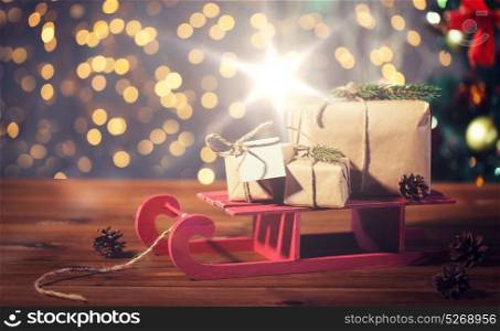 holidays, presents, new year and celebration concept - close up of gift boxes with blank note on red wooden sleigh over christmas tree and lights background. close up of christmas gift boxes on wooden sleigh