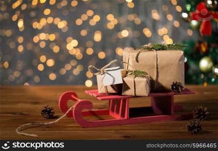 holidays, presents, new year and celebration concept - close up of gift boxes with blank note on red wooden sleigh over christmas tree and lights background