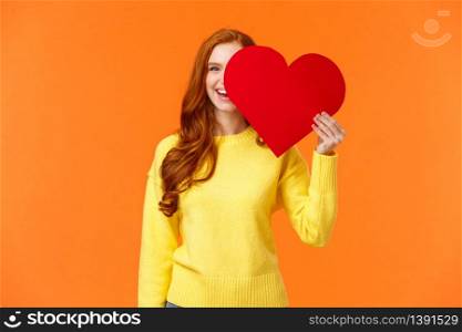 Holidays, romance concept. Cheerful girl showing affection, give heart to girlfriend, hiding half face behind valentines card, smiling joyfully, prepare romantic gesture for date, orange background.. Holidays, romance concept. Cheerful girl showing affection, give heart to girlfriend, hiding half face behind valentines card, smiling joyfully, prepare romantic gesture for date, orange background