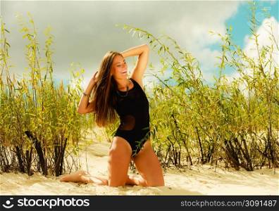 Holidays, summer and beauty concept. Sensual girl in black swimwear posing on sandy beach, grassy dunes. Pretty woman on the sea coast.
