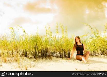 Holidays, summer and beauty concept. Sensual girl in black swimwear posing on sandy beach, grassy dunes. Pretty woman on the sea coast.