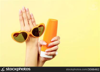 Holidays summer fashion eyes protection and skin care concept. Woman holding in hand heart shaped sunglasses sunscreen lotion, yellow background