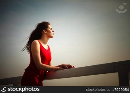 Holidays summer relaxation concept. Young thoughtful woman wearing red dress relaxing on pier outdoor