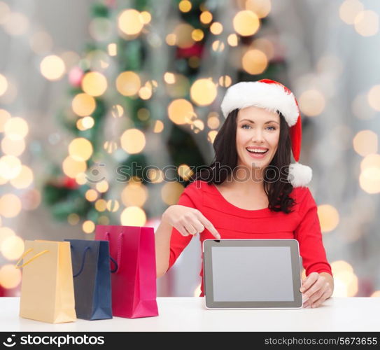 holidays, technology, advertising and people concept - smiling woman in santa helper hat with shopping bags and tablet pc computer over christmas tree background