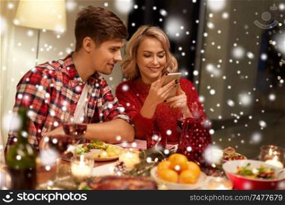 holidays, technology and celebration concept - happy couple having christmas dinner at home and using smartphone over snow. couple with smartphone at home christmas dinner