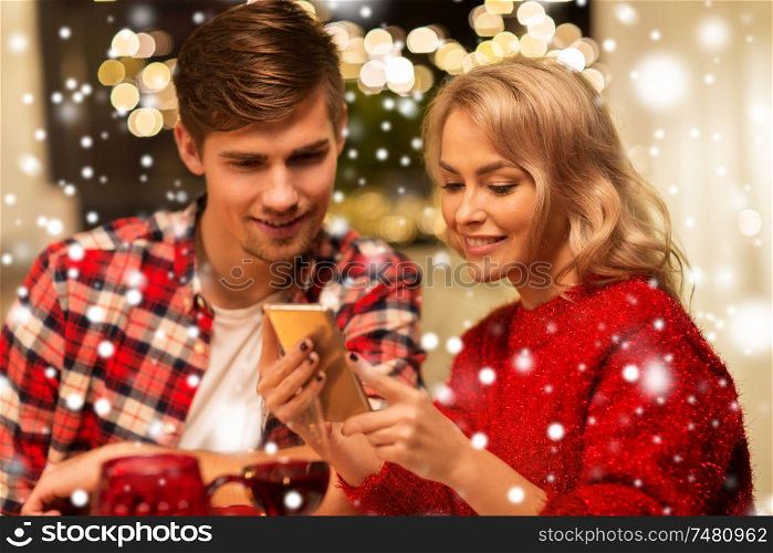 holidays, technology and celebration concept - happy couple having christmas dinner at home and using smartphone over snow. couple with smartphone at home christmas dinner