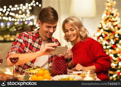 holidays, technology and celebration concept - happy couple having christmas dinner at home and photographing food by smartphone. couple with smartphone at home christmas dinner