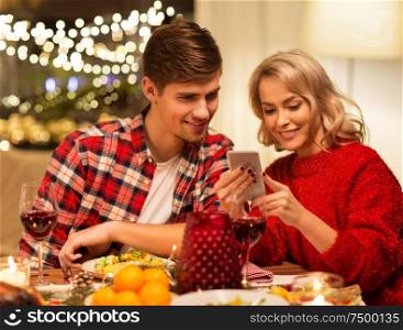 holidays, technology and celebration concept - happy couple having christmas dinner at home and using smartphone. couple with smartphone at home christmas dinner