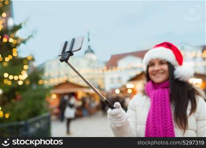 holidays, technology and people concept - happy woman taking picture with smartphone selfie stick at old town hall square in tallinn. woman taking selfie with smartphone at christmas
