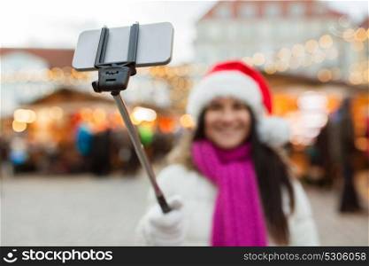 holidays, technology and people concept - happy woman taking picture with smartphone selfie stick at christmas market at old town hall square in tallinn. woman taking selfie by smartphone at christmas