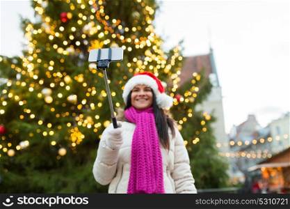 holidays, technology and people concept - happy woman taking picture with smartphone selfie stick at christmas tree at old town hall square in tallinn. woman taking selfie with smartphone at christmas