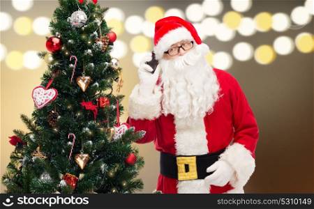 holidays, technology and people concept - man in costume of santa claus and christmas tree calling on smartphone. santa claus calling on smartphone at christmas