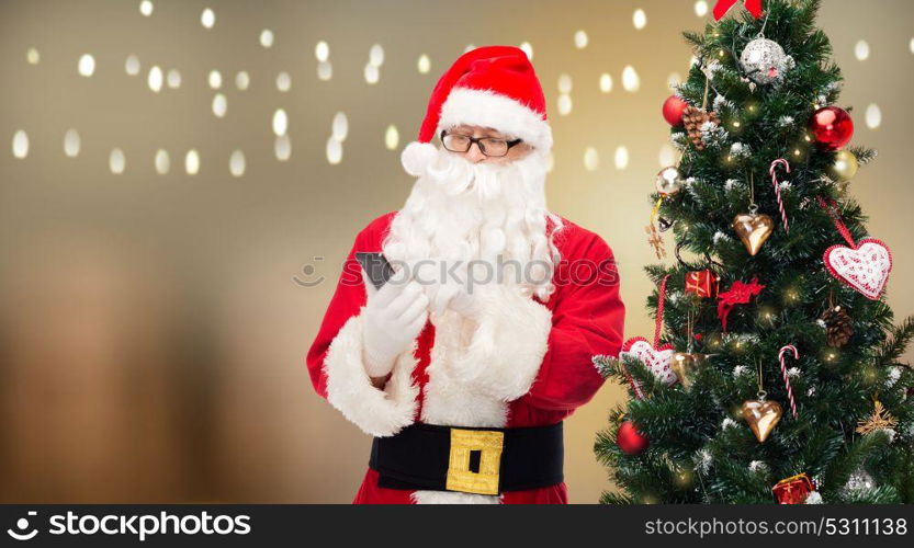 holidays, technology and people concept - man in costume of santa claus and christmas tree dialing number on smartphone. santa claus with smartphone at christmas tree