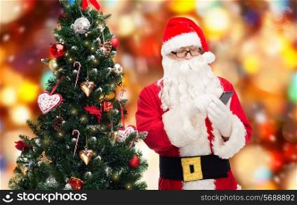 holidays, technology and people concept - man in costume of santa claus with smartphone and christmas tree over red lights background