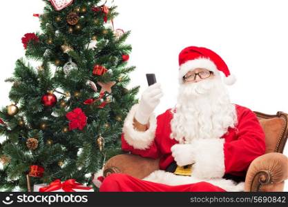 holidays, technology and people concept - man in costume of santa claus with smartphone, presents and christmas tree