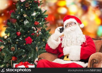 holidays, technology and people concept - man in costume of santa claus with smartphone, presents and christmas tree over red lights background