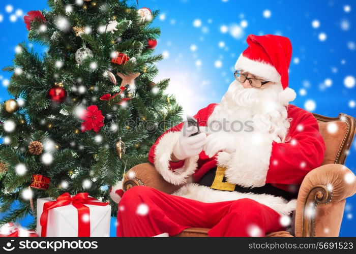 holidays, technology and people concept - man in costume of santa claus with smartphone, presents and christmas tree over blue snowy background