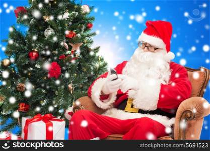 holidays, technology and people concept - man in costume of santa claus with smartphone, presents and christmas tree over blue snowy background