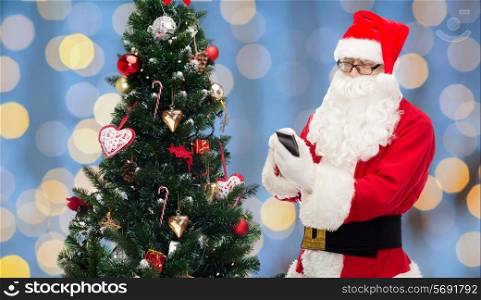 holidays, technology and people concept - man in costume of santa claus with smartphone and christmas tree over blue lights background
