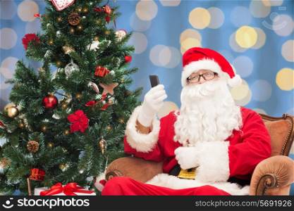 holidays, technology and people concept - man in costume of santa claus with smartphone, presents and christmas tree over blue lights background