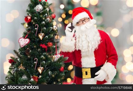 holidays, technology and people concept - man in costume of santa claus with smartphone and christmas tree over lights background