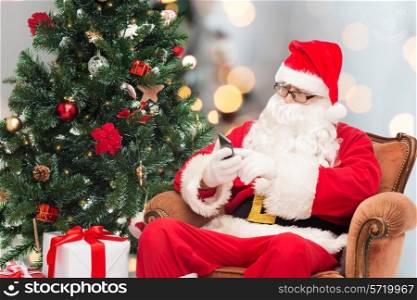 holidays, technology and people concept - man in costume of santa claus with smartphone, presents and christmas tree sitting in armchair over lights background