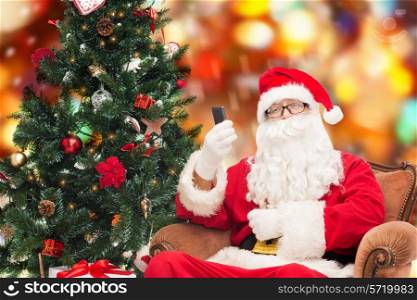 holidays, technology and people concept - man in costume of santa claus with smartphone, presents and christmas tree over red lights background