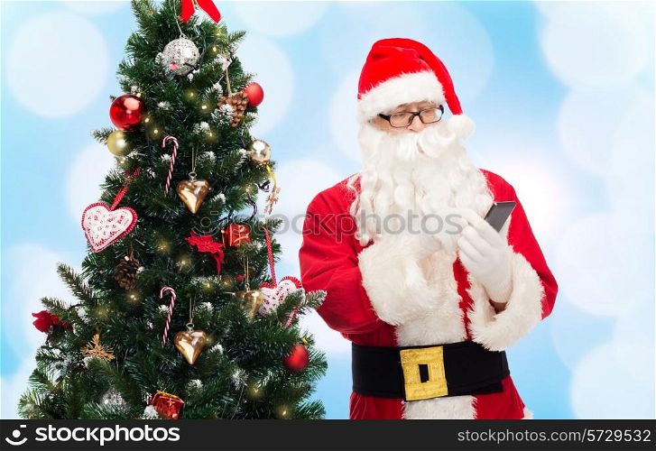 holidays, technology and people concept - man in costume of santa claus with smartphone and christmas tree over blue lights background