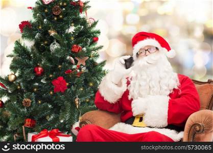 holidays, technology and people concept - man in costume of santa claus with smartphone, presents and christmas tree over lights background