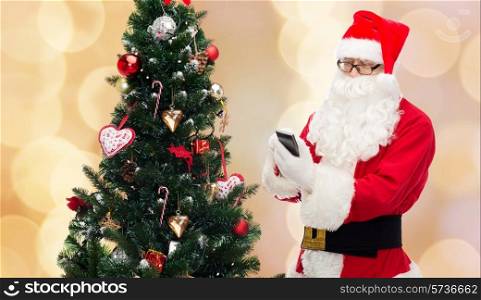holidays, technology and people concept - man in costume of santa claus with smartphone, presents and christmas tree over beige lights background