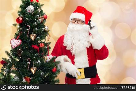 holidays, technology and people concept - man in costume of santa claus with smartphone and christmas tree over beige lights background