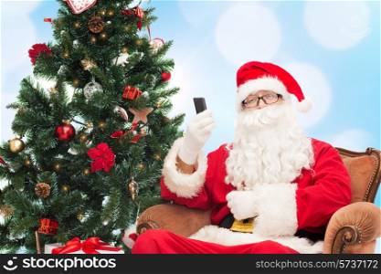holidays, technology and people concept - man in costume of santa claus with smartphone, presents and christmas tree over blue lights background