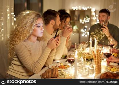 holidays, technology and people concept - sad young woman with smartphone at christmas dinner party with friends at home. woman with smartphone at dinner party with friends