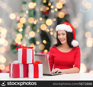 holidays, technology and people concept - smiling woman in santa helper hat with gift boxes and laptop computer over christmas tree with lights background