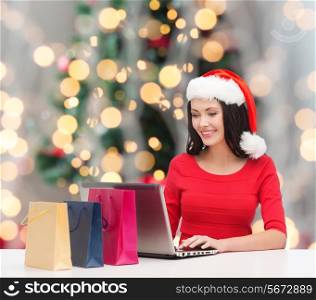 holidays, technology and people concept - smiling woman in santa helper hat with shopping bags and laptop computer over christmas tree with lights background
