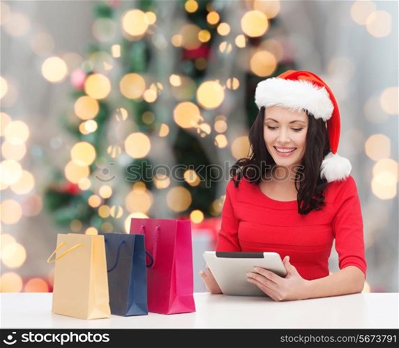 holidays, technology and people concept - smiling woman in santa helper hat with shopping bags and tablet pc computer over christmas tree with lights background