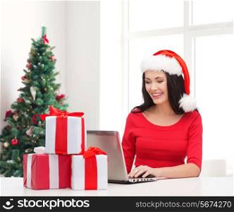 holidays, technology and people concept - smiling woman in santa helper hat with gift boxes and laptop computer over living room with christmas tree background