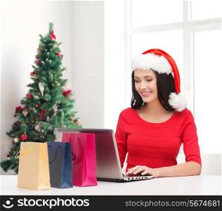 holidays, technology and people concept - smiling woman in santa helper hat with shopping bags and laptop computer over living room with christmas tree background