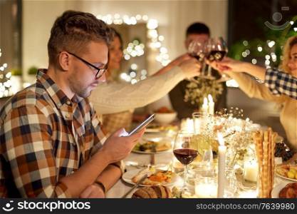 holidays, technology and people concept - young man in glasses with smartphone at christmas dinner party with friends at home. man with smartphone at dinner party with friends