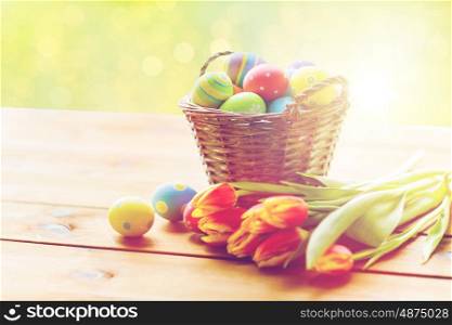 holidays, tradition and object concept - close up of colored easter eggs in basket and tulip flowers on wooden table over green lights background