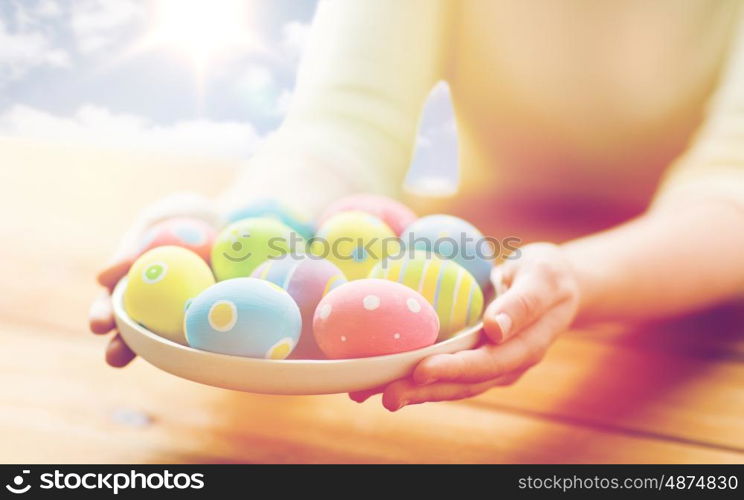 holidays, tradition and people concept - close up of woman hands holding colored easter eggs on plate over blue sky and sun background