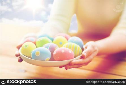 holidays, tradition and people concept - close up of woman hands holding colored easter eggs on plate over blue sky and sun background