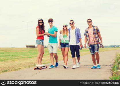 holidays, vacation, love and friendship concept - group of smiling teenagers walking and riding on skateboards outdoors