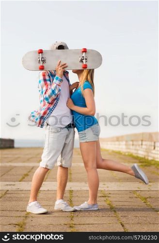 holidays, vacation, love and people concept - couple kissing and hiding their faces behind skateboard outdoors